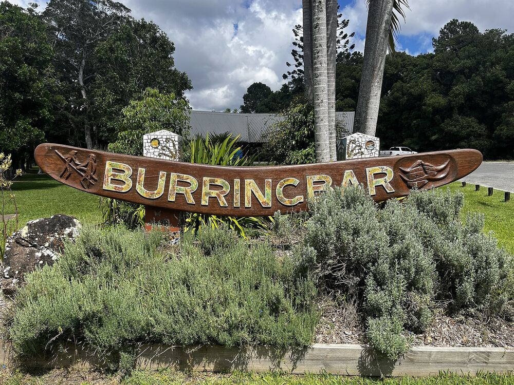 The carved wooden Burringbar village sign in a bed of rosemary, with palms behind